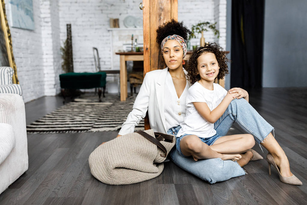 Uma menina encaracolado bonito e sua mãe estão sentados na sala de estar no chão
 - Foto, Imagem