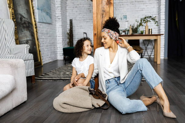 A cute curly girl and her mother are sitting in the living room on the floor - Photo, Image