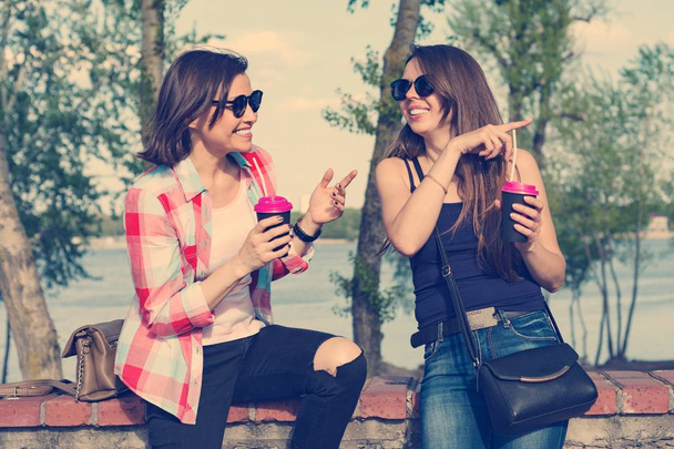 Happy two female friends drinking coffee and having fun. Background nature, park, river. Urban lifestyle and friendship concept - Zdjęcie, obraz