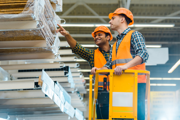 smiling indian warehouse worker pointing with finger at construction materials while standing on scissor lift near colleague  - Photo, Image