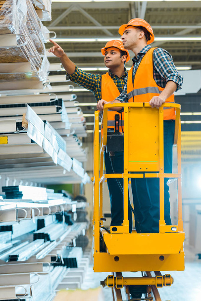 serious indian worker pointing with finger at construction materials while standing on scissor lift near colleague  - Photo, Image