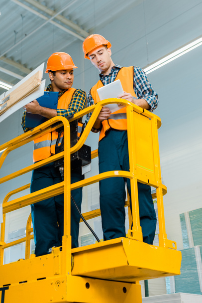 concentrated multicultural workers with digital tablet and clipboard standing on scissor lift - Photo, Image
