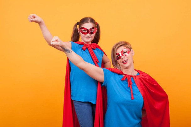 Beautiful mother and her little daughter dressed like superheros are keeping one hand up over yellow background - Photo, Image