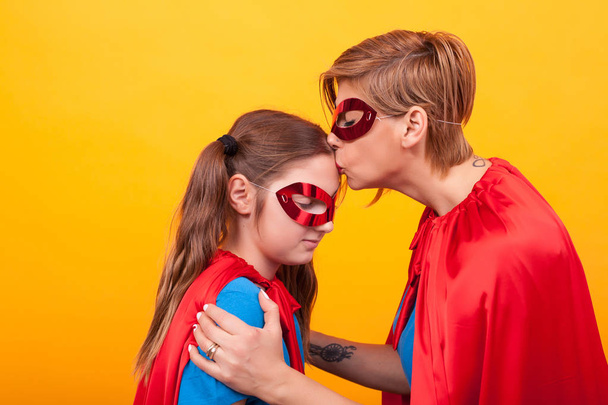 Мать в костюме супермена целует девочку в лоб на жёлтом фоне.
 - Фото, изображение
