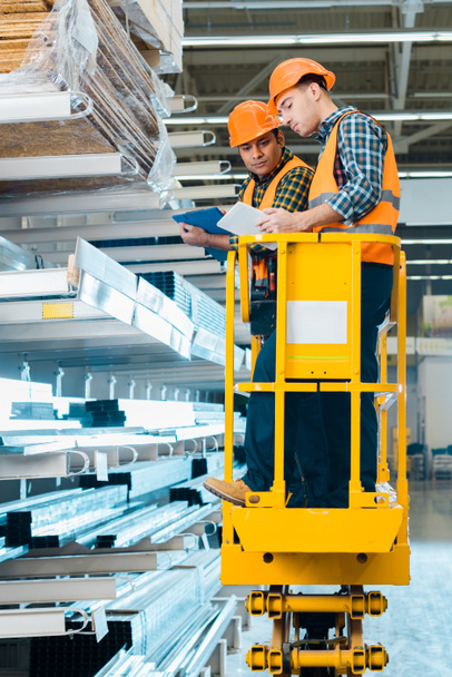 multicultural workers with digital tablet and clipboard standing on scissor lift near shelves with metallic construction materials - Photo, Image