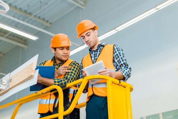 thoughtful multicultural workers with digital tablet and clipboard standing on scissor lift - Photo, Image