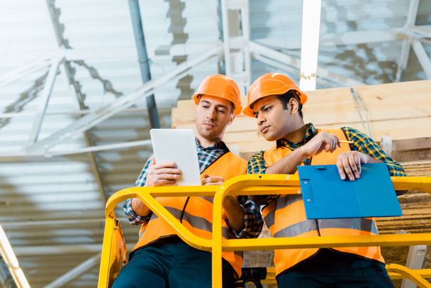 thoughtful multicultural workers using digital tablet while standing on scissor lift in warehouse - Photo, Image