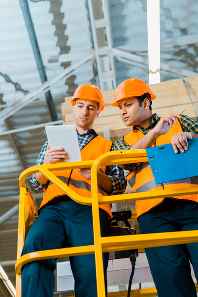 serious multicultural warehouse workers using digital tablet while standing on scissor lift  - Foto, imagen