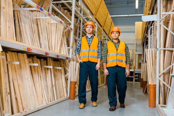 multicultural warehouse workers in safety vasts and helmets walking along racks with wooden construction materials - Photo, Image