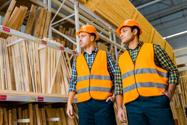 мультикультурные складские работники в защитных касках и шлемах, стоящих рядом со стойками с деревянными строительными материалами
 - Фото, изображение