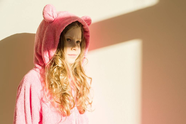 Portrait of a girl in soft warm pink pajamas. Girl 7 years old, blonde with long curly hair, in a hood, looking away. Background light house wall, solar lighting from the window, copy space - Photo, image