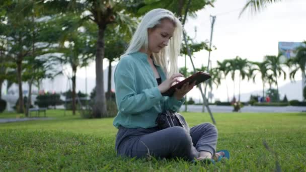 Όμορφη λεπτή γυναίκα με μακριά ξανθά μαλλιά σε πράσινο πουκάμισο κάθεται στο έδαφος και χρησιμοποιώντας το smartphone πάνω από το παρασκήνιο το πάρκο. Κορίτσι στην πλατεία αγγίζοντας την οθόνη και το χαμόγελο. - Πλάνα, βίντεο