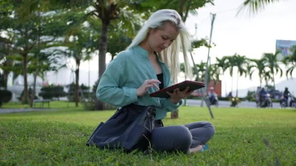 Gyönyörű karcsú nő, hosszú szőke haja zöld inget ül a földön, és segítségével okostelefon háttérben a parkban. Lány a téren megható képernyő és a mosoly. - Felvétel, videó