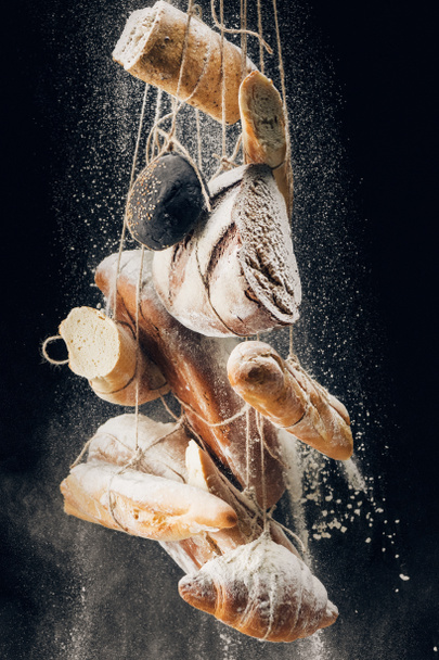 white flour falling at bread, baguettes and croissant on ropes on black background - Fotó, kép