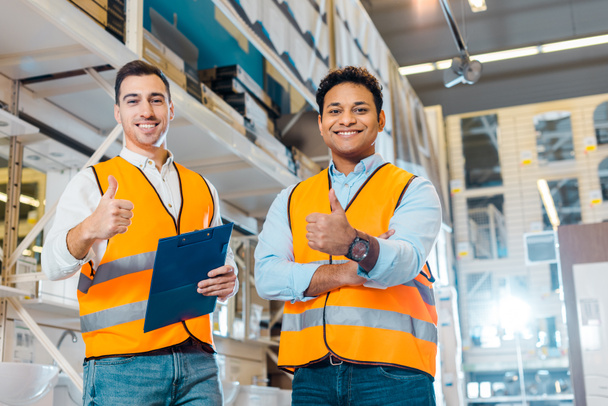 cheerful multicultural warehouse workers showing thumbs up and looking at camera - Photo, Image