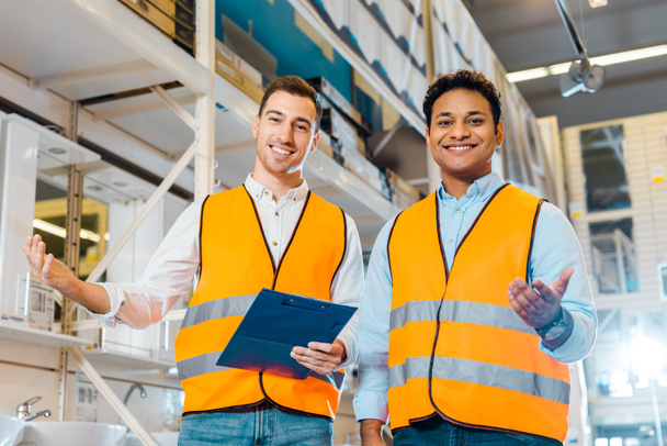 smiling multicultural warehouse workers in safety vests smiling and looking at camera - 写真・画像