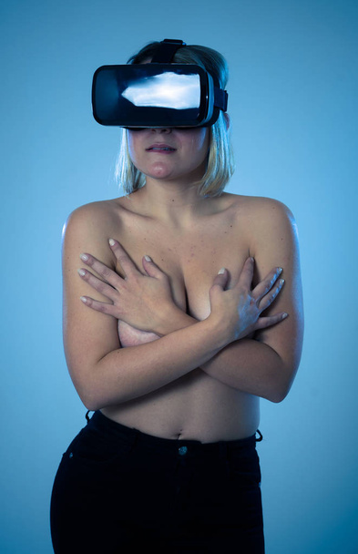 Sensual attractive woman using augmented or virtual reality goggles feeling excited exploring virtual sexuality while getting topless covering her breast. In cyber sex VR simulator and new technology. - 写真・画像