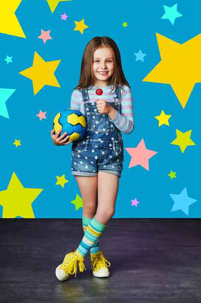 Cute little girl in denim against blue wall. Fashion concept.Child model in jeans overalls posing in studio, multi-colored stars background . - Foto, immagini