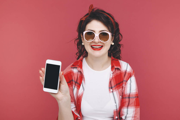 Portrait of an excited beautiful girl wearing red shirt, holding mobile phone isolated over pink background. - Photo, Image