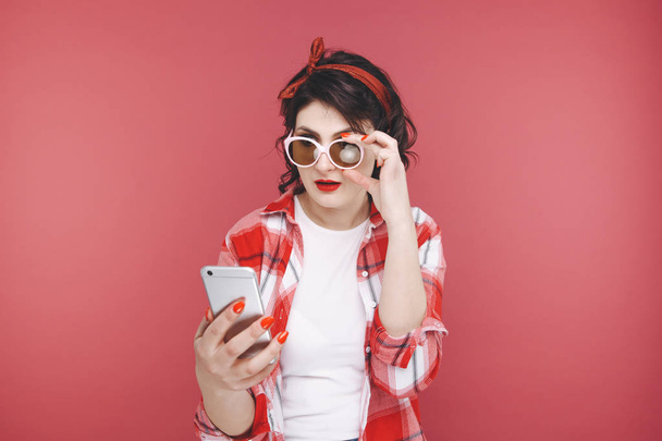 Retrato de una hermosa chica excitada usando camisa roja, sosteniendo el teléfono móvil aislado sobre fondo rosa
. - Foto, Imagen