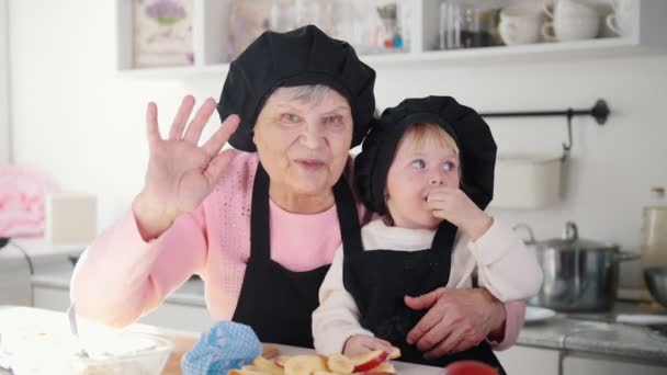 Een oude vrouw met een klein meisje zwaaiende handen en kijken in de camera in de keuken. - Video