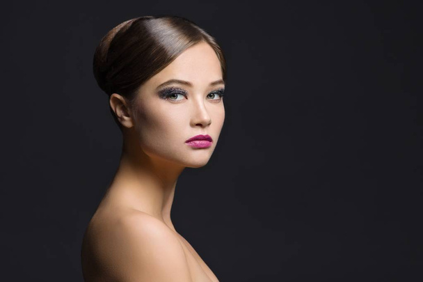 Close-up portrait of young brunette girl with brushed hair, evening make-up, naked shoulders and long neck. Woman looking at camera, black background, copy space - Photo, image