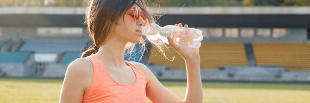 Young girl teenager drinking water from bottle after running at stadium, panoramic banner on sunny spring evening - Foto, Imagem