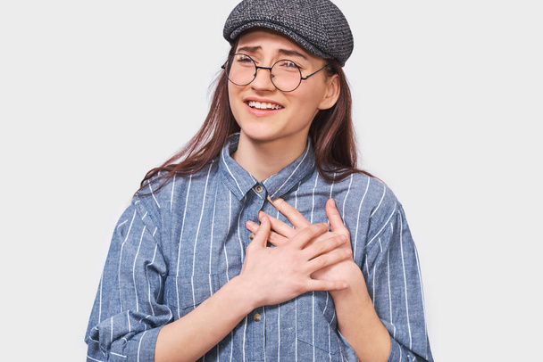 Portrait of kind grateful young woman smiling and keeps both palms on chest, wears eyewear, dressed in blue striped t-shirt, isolated over white background. People, emotions, feeling and body language - Photo, Image