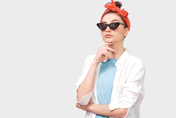 Beautiful serious brunette young woman wears black sunglasses, white shirt and trendy red headband, looks seriously directly into camera, poses against white studio background. People emotions concept - Photo, Image