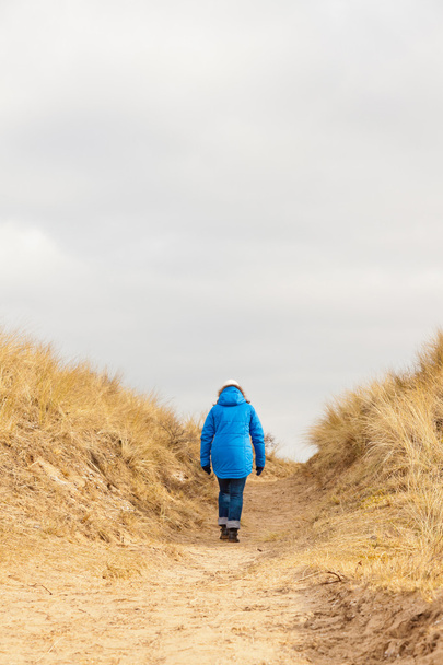 Turista caminando por sendero en paisaje de dunas cubiertas de hierba con cielo nublado
 - Foto, Imagen