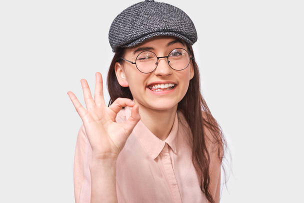 Ok のジェスチャーを示すトレンディな灰色キャップと丸い透明なメガネを着て満足している若い女性のスタジオ画像を閉じます。かなり学生女の子笑顔といい兆しを見せ. - 写真・画像