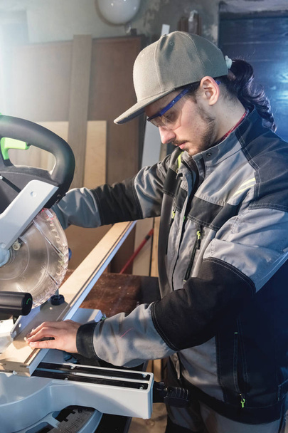Young brunette man wearing a cap in a gray jacket by profession a carpenter cuts wooden boards with a circular saw on a workbench table in a workshop. Professional equipment in home workshops - Foto, afbeelding