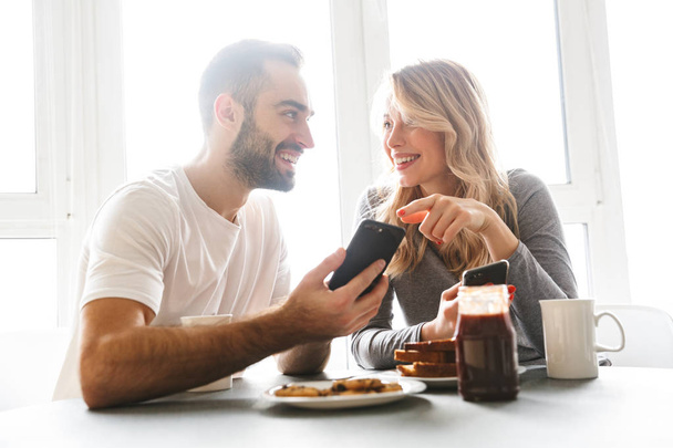 Loving couple sitting at the kitchen have a breakfast using mobile phones. - Foto, Imagem