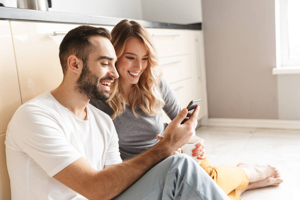 Amazing young loving couple sitting at the kitchen using mobile phone on a floor. - 写真・画像