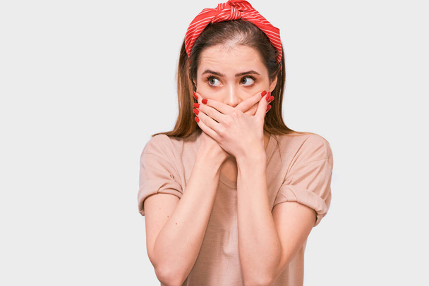 Portrait of shocked horrified young woman covering mouth with hands feel scared looking away, isolated on white studio wall. Pretty female with amazed face afraid in silence weraing red headband. - Photo, Image