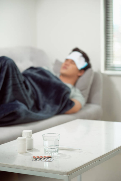 sick wasted man lying in sofa suffering cold and winter flu virus having medicine tablets in health care concept looking temperature on thermometer - Fotoğraf, Görsel