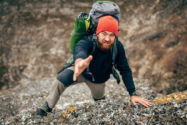 Horizontal image of traveler bearded man trekking and mountaineering during his journey. Young tourist male holds out his hand for help during hiking in mountains. Travel, people, sport, lifestyle - Фото, изображение
