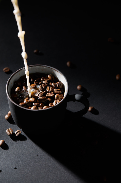 Xícara de café com grãos de café e derramando leite na superfície escura
 - Foto, Imagem