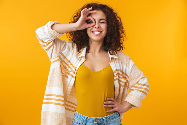 Beautiful young curly girl posing isolated over yellow wall background with okay gesture. - Photo, image
