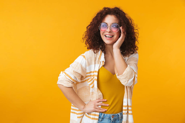 Beautiful young curly girl posing isolated over yellow wall background wearing sunglasses. - Фото, изображение