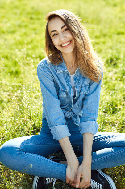 portrait of attractive woman in jeans sitting in a park on the grass at sunny summer day. - Fotografie, Obrázek