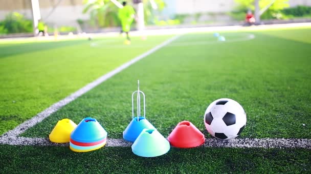 Fußball und Markierungskegel stehen auf grünem Kunstrasen mit verschwommenen Kinderfußballern beim Training, Fußballtrainingsgeräte in der Fußballakademie. - Filmmaterial, Video