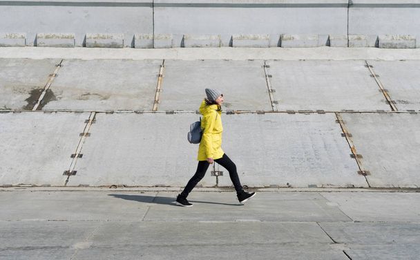 Profile view: a girl in a yellow jacket walks on a walk in wide strides on a concrete surface. - Foto, Imagem