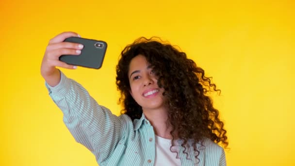 Beautiful woman with curly hair holding and using smart phone to film herself in yellow studio. Female using technology networking on holiday, lifestyle. 4k. - Video, Çekim