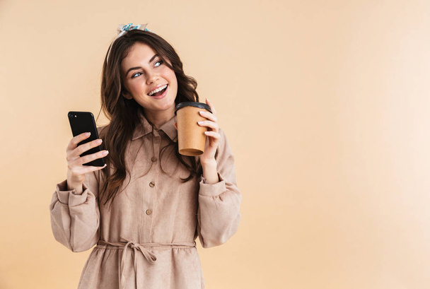 Joven bonita mujer posando aislada sobre fondo beige pared celebración de teléfono móvil charlando beber café
. - Foto, Imagen