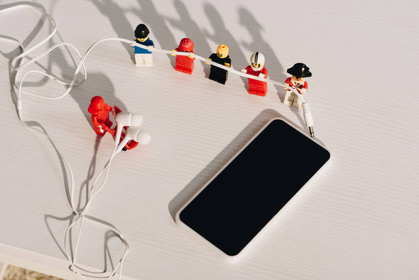 KYIV, UKRAINE - MARCH 15, 2019: lego figurines holding earphones near smartphone with blank screen on white table - Photo, Image