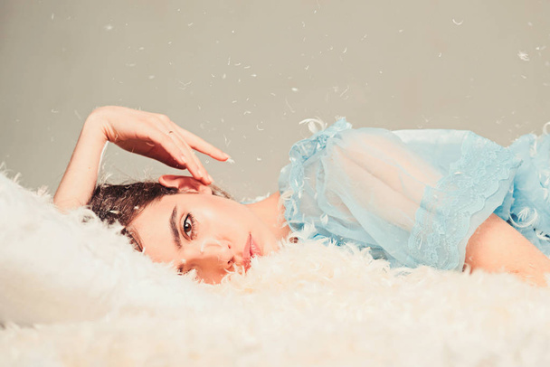Girl on calm face lay on bed covered with feathers and fluff. Soft and gentle concept. Lady in transparent blue nightie lay on bed, grey background. Woman with long hair in tender pajama relaxing. - Foto, afbeelding