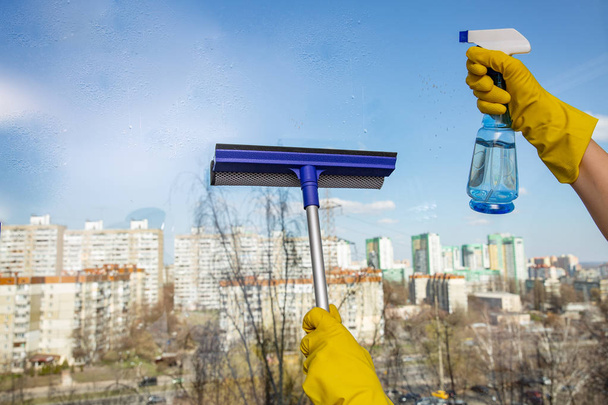 Manos de mujer con guantes de goma amarillos que sostienen la herramienta de limpieza de vidrio de ventana y botella de spray con detergente sobre el fondo de la ventana
 - Foto, imagen