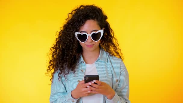 Young female smiling and using mobile phone over yellow background. Beautiful mixed race girl holding and texting with smart phone. Woman with technology. - Materiał filmowy, wideo