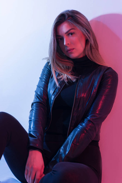 Seksi genç kadın portre, renkli arka plan üzerinde. Baştan çıkarıcı esmer modeli kız siyah ceketli. Kız oturuyor. Kırmızı ve mavi ışık. Gece hayatı. Yakın çekim - Fotoğraf, Görsel
